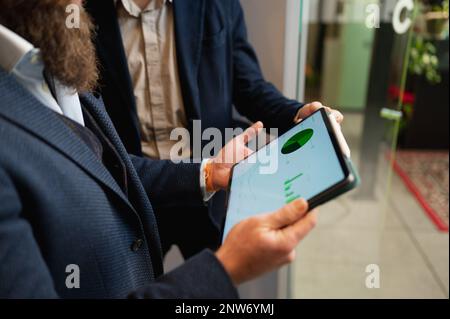 Unbekannte Geschäftsmänner in eleganten Anzügen, die ein Tablet halten und Diagramme analysieren. Stockfoto