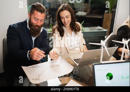 Ein paar junge Kollegen arbeiten mit fröhlichen positiven Gesichtern zusammen und sitzen am Schreibtisch im Büro. Geschäftsleute analysieren Diagramme und verwenden Comput Stockfoto