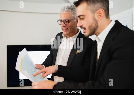 Ein junger Geschäftsmann bittet einen leitenden Kollegen um Rat zu einer Strategie. Geschäftsleute am Arbeitsplatz, die Diagramme oder Entwürfe halten und analysieren. Stockfoto