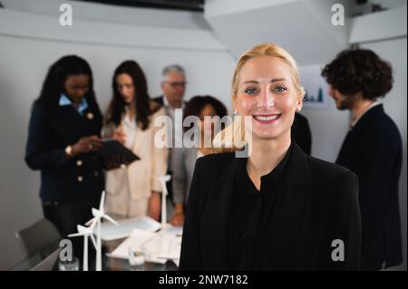 Porträt einer jungen, attraktiven blonden Geschäftsfrau, die steht und lächelt. Unscharfe Teamarbeit am Arbeitsplatz. Stockfoto