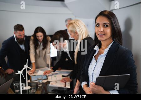 Porträt einer jungen arabischen Geschäftsfrau, die ein digitales Tablet hält und in die Kamera schaut. Team arbeitet an Hintergrundinformationen. Stockfoto