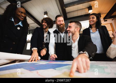Glückliche Teamarbeit mit unterschiedlicher ethnischer Zugehörigkeit, die die Zusammenarbeit genießt. Pov-Mann zeigt etwas auf Blaupausen auf dem Schreibtisch. Stockfoto