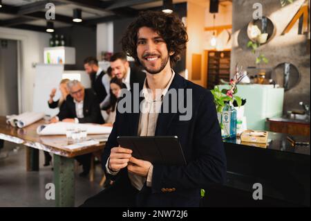 Junger Hipster, der vor der Kamera posiert, lächelt und ein digitales Tablet hält. Multiethnische Geschäftsleute, die am Arbeitsplatz im Hintergrund arbeiten Stockfoto