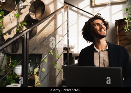 Junger Hipster, der auf einer Treppe einer Lounge sitzt, mit einem Laptop und mit positivem Gesicht aus dem Fenster schaut. Kreativität und Konzept für kleine Unternehmen Stockfoto