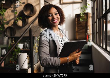 Porträt einer lächelnden lateinischen Frau in eleganter Kleidung, die ein digitales Tablet hält und die Kamera lächelt. Stockfoto