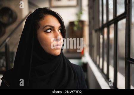 Nahaufnahme einer jungen attraktiven araberin mit schwarzem Schleier. Weibliches Profil, das aus dem Fenster schaut, mit ernstem Gesicht. Stockfoto