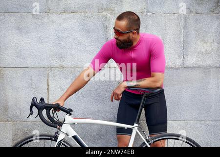 Ein bärtiger Radfahrer in Brille mit einem Fahrrad auf dem Hintergrund einer grauen Mauer in der städtischen Landschaft, eine tägliche Fahrradtour. Stockfoto