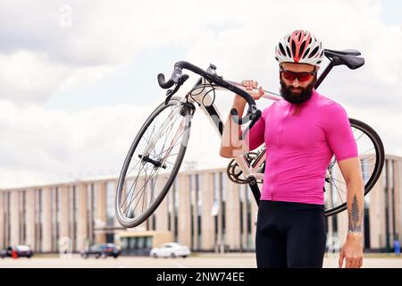 Bärtiger männlicher Radfahrer mit Brille und ein Helm schaut auf die Kamera mit einem Fahrrad in der Hand gegen den blauen Himmel, Lifestyle, Cardio-Training Stockfoto