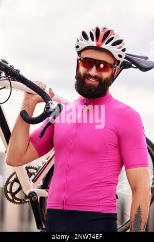 Bärtiger männlicher Radfahrer mit Brille und ein Helm schaut auf die Kamera mit einem Fahrrad in der Hand gegen den blauen Himmel, Lifestyle, Cardio-Training Stockfoto