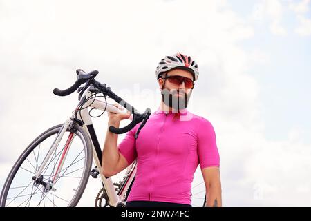 Bärtiger männlicher Radfahrer mit Brille und ein Helm schaut auf die Kamera mit einem Fahrrad in der Hand gegen den blauen Himmel, Lifestyle, Cardio-Training. Stockfoto