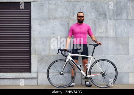 Ein bärtiger Radfahrer in Brille mit einem Fahrrad auf dem Hintergrund einer grauen Mauer in der städtischen Landschaft, eine tägliche Fahrradtour. Stockfoto