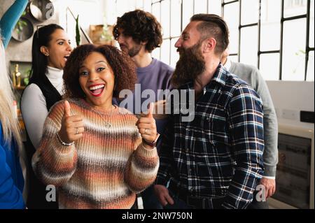 Hispanische Frau, die mit erhobenem Daumen in die Kamera schaut. Junge gemischte Rassen versammeln sich im Hintergrund. Stockfoto