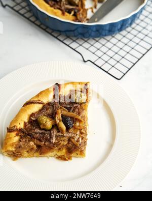 Pissaladiere Zwiebelkuchen mit Sardellen und schwarzen Oliven. Das abgeschnittene Stück des Tortendiagramms liegt auf der Platte, der verbleibende Tortendiagramm im Werkzeug im Hintergrund Stockfoto