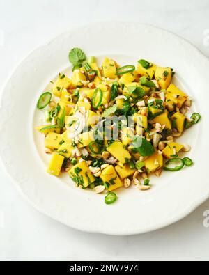 Thailändischer Mangosalat auf weißem Teller auf weißem Hintergrund. Salat aus Mango mit Erdnüssen, Chili und Fischsauce, dekoriert mit Minzblättern. Stockfoto