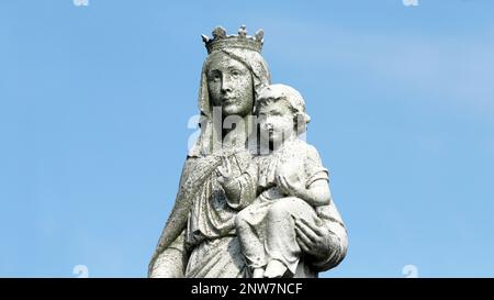 1920er Statue der Jungfrau Maria mit Jesuskind auf dem Friedhof. Getragen, verwittert und bedeckt mit Moos am blauen Himmel Stockfoto