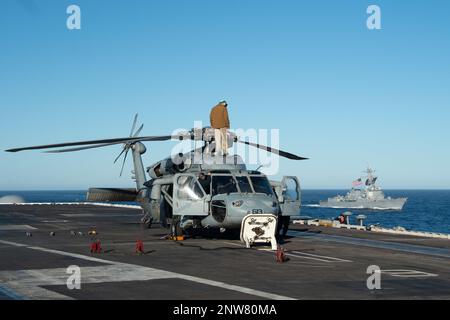 230207-N-PQ495-1273 PAZIFIK (7. Februar 2023) Seeleute an Bord des Flugzeugträgers USS Carl Vinson der Nimitz-Klasse (CVN 70) führen Wartungsarbeiten an einem MH-60s Sea Hawk durch, der an den "Schwarzen Rittern" des Helicopter Sea Combat Squadron (HSC) 4 befestigt ist, während Arleigh Burke-Klasse Guided-Missile Destroyer USS Sterett (DDG 104) neben DDG fährt. Vinson führt derzeit routinemäßige Seeeinsätze durch. Stockfoto