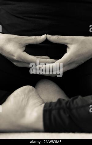 Vertikales BW-Detail von Händen und Füßen einer jungen Frau, die in Innenräumen meditiert. Yogini macht eine kontemplative Handgeste Dhyana Mudra Stockfoto