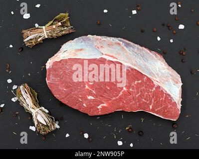 Frisches rohes Rumpffleisch, das von der Picanha-Kappe auf einer schwarzen Schieferplatte geschnitten wurde, mit Salz, Pfeffer und Gewürzbündel Stockfoto