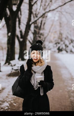 Eine junge Frau, die warme Winterkleidung trägt und eine Strickmütze trägt, lächelt fröhlich, wenn sie im Schnee steht und mit ihrem Handy einen Kaffeebecher hält Stockfoto