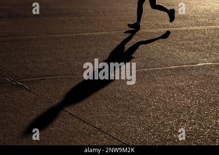 Beine eines unbekannten männlichen Joggers, der bei Sonnenuntergang in der Stadt läuft, mit seinem langen Schatten, der auf einen dunklen Bürgersteig projiziert wird Stockfoto