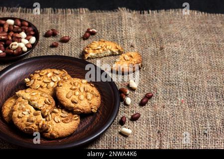 Hausgemachte Erdnusskekse auf einem braunen Teller mit rohen Erdnüssen im Hintergrund. Rustikales Essen. Speicherplatz kopieren Stockfoto