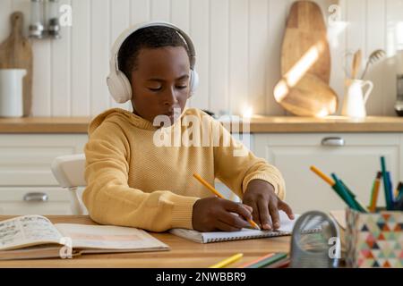 Ein durchdachter Schuljunge mit Ohrhörern aus verschiedenen Rassen macht Sport in Lehrbuchschriften mit Bleistiften, die zu Hause sitzen Stockfoto