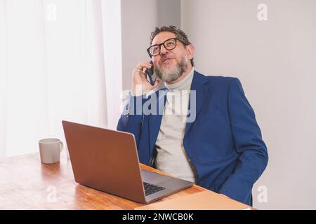 Ein erwachsener Geschäftsmann mit Bart und Brille, der am Telefon telefoniert, während er mit einem Laptop am Schreibtisch sitzt. Er trägt einen weißen Rollkragenpullover und Bl Stockfoto
