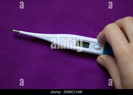 Weibliche Hand hält ein elektronisches Thermometer auf violettem Hintergrund, Medizin und Pharmazie, Gesundheit und Behandlung, hohe Temperatur Stockfoto