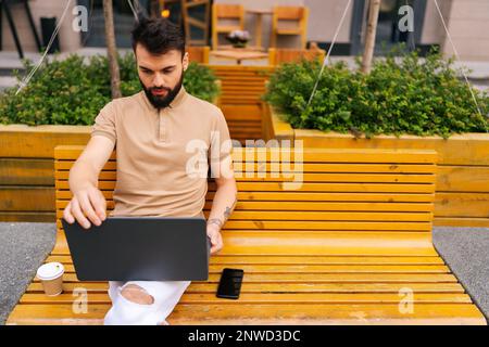 Blick aus dem großen Blickwinkel auf einen fokussierten, bärtigen jungen Mann, der per Fernzugriff an einem Laptop arbeitet, auf einer Bank auf der Straße der Stadt sitzt und Kaffee zum Mitnehmen mitbringt. Stockfoto