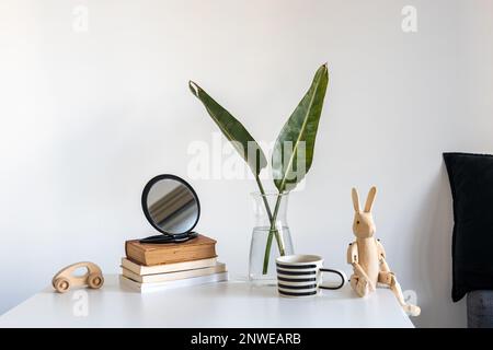 Stillleben mit einem Holzhasen auf Scharnieren im Innenraum. Eine Vase mit zwei grünen Palmenfronten, einem weiß-schwarz gestreiften Becher, einem Stapel Bücher und einem Woo Stockfoto