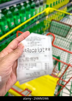 Hand halten und einen Kaufbeleg mit Supermarktwagen im Hintergrund zeigen. Einkaufskosten und Inflationskonzept. Stockfoto