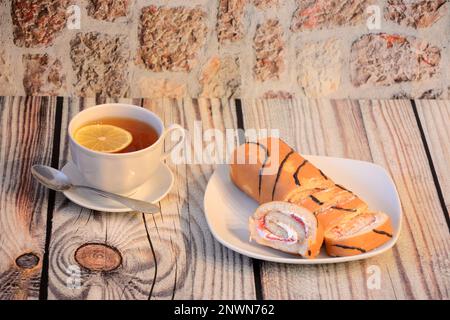 Eine Tasse heißen Tee mit einer Scheibe Zitrone und ein Teller mit Keksrollen mit Proteincreme auf einem Holztisch. Nahaufnahme. Stockfoto