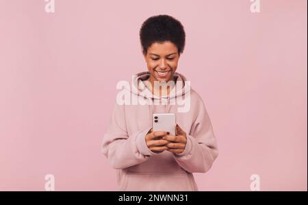 Fröhliche junge afrikanische Frau, die eine Nachricht auf ihrem Handy tippt, während sie über einem rosa Hintergrund steht Stockfoto