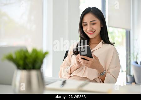 Charmante asiatische Millennials-Geschäftsfrau, die ihr Smartphone an ihrem Schreibtisch in einem modernen Büro verwendet. Chat, SMS, E-Mail Stockfoto