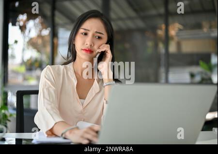 Gestresste und verwirrte asiatische Millennials-Geschäftsfrau telefoniert mit einem Geschäftskunden an ihrem Schreibtisch und hat einen ernsthaften Anruf. Stockfoto