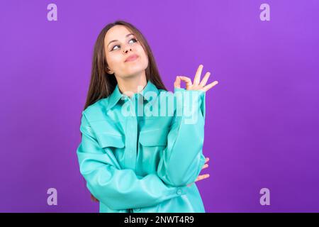 Schöne Frau in lässiger Kleidung, die mit der Hand ein Zeichen für "OK" macht. Erfolgreicher Ausdruck, violetter Hintergrund Stockfoto