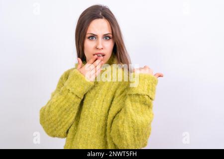 Eine attraktive Frau, die mit dem Finger auf den Textbereich auf weißem Hintergrund zeigt, ein grüner Wollpullover, überrascht Stockfoto