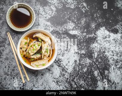 Asiatische Knödel in Brühe, Schüssel, Essstäbchen, Sojasauce, rustikaler Steinhintergrund. Draufsicht. Platz für Text. Chinesische Knödel zum Abendessen. Nahaufnahme. Stockfoto