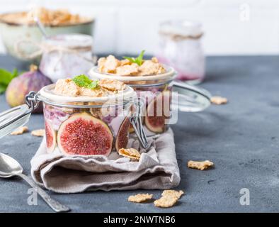 Hausgemachter gesunder Joghurt in Glastopf mit Müsli, Feigen, Minze auf rustikalem Betonhintergrund. Gesundes Frühstück am Morgen. Frisch zubereiteter Joghurt mit Stockfoto