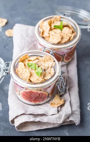 Hausgemachter gesunder Joghurt in Glastopf mit Müsli, Feigen, Minze auf rustikalem Betonhintergrund. Gesundes Frühstück am Morgen. Frisch zubereiteter Joghurt mit Stockfoto