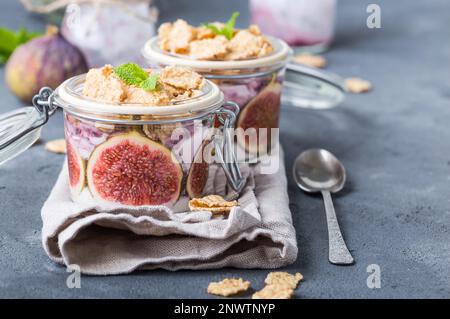 Hausgemachter gesunder Joghurt in Glastopf mit Müsli, Feigen, Minze auf rustikalem Betonhintergrund. Gesundes Frühstück am Morgen. Frisch zubereiteter Joghurt mit Stockfoto