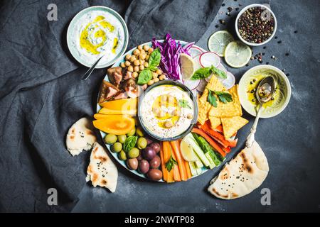 Meze-Platte mit Hummus, Joghurtdip und verschiedenen Snacks. Hummus in der Schüssel, Gemüsestangen, Kichererbsen, Oliven, Pita, Chips. Platte mit Mitte Stockfoto