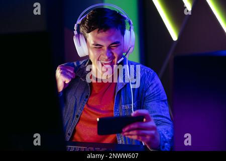 Männlicher Videospielspieler jubelt und feiert, während er eine Live-Übertragung eines Spiels auf seinem Smartphone streamt. Ein junger Mann, der sich einem Online-Publikum in wa anschließt Stockfoto
