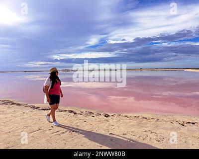 Lateinische Erwachsene Frau mit Shorts, pinkfarbenem Hemd, Hut und Sonnenbrille wandert auf dem Sand neben der pinkfarbenen Lagune Las Coloradas in Yucatan Mexico Stockfoto