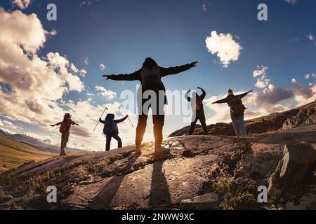 Die Silhouetten einer Gruppe von Wanderer stehen mit offenen Armen in siegreichen Posen vor der Sonne und den Bergen bei Sonnenuntergang Stockfoto
