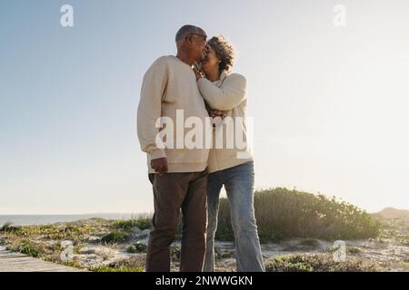 Seniorin flüstert ihrem Mann zu, während sie am Strand eine hölzerne Fußbrücke hinuntergeht. Glückliches älteres Paar, das danach einen erfrischenden Urlaub genießt Stockfoto