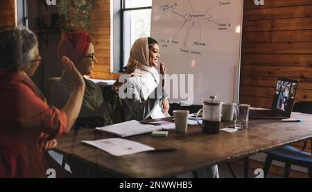 Eine Gruppe glücklicher Geschäftsfrauen, die während eines Online-Meetings in einem modernen Büro mit der Hand winken. Multikulturelle Geschäftsfrauen, die eine Videokonferenz veranstalten Stockfoto