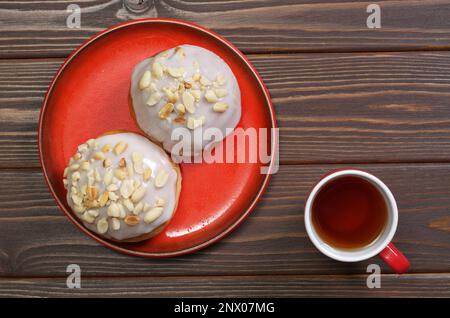 Zwei glasierte Brötchen mit Erdnüssen auf rotem Teller und eine Tasse Tee auf einem Holztisch, Blick von oben Stockfoto