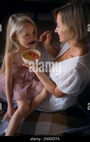 Blonde Mutter und Tochter mit langen Haaren umarmen und küssen sich. Mutters Liebe, Zärtlichkeit und Fürsorge. Die Tochter ist beleidigt und traurig, beleidigt. Eine Frau, die ein Kind zu Hause tröstet. Hochwertiges Foto Stockfoto