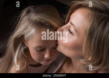 Blonde Mutter und Tochter mit langen Haaren umarmen und küssen sich. Mutters Liebe, Zärtlichkeit und Fürsorge. Die Tochter ist beleidigt und traurig, beleidigt. Eine Frau, die ein Kind zu Hause tröstet. Hochwertiges Foto Stockfoto